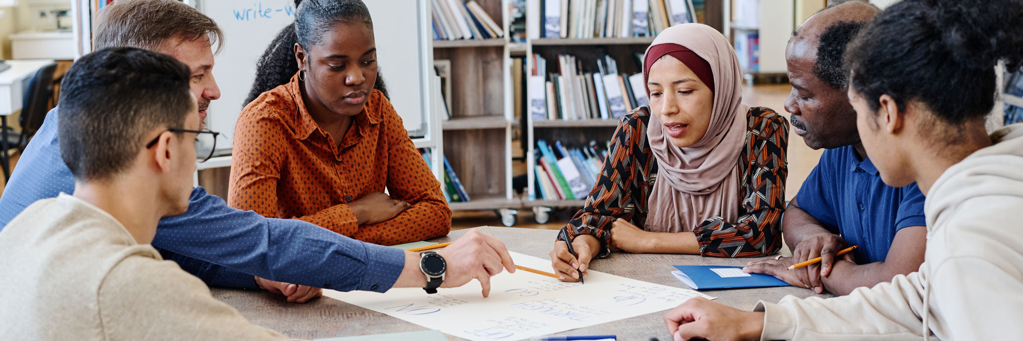 a diverse group of adults sitting around a table in a community library working together on paperwork